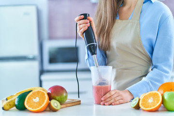 Woman in apron using hand blender for preparing a fresh organic fruit smoothie at home in the...