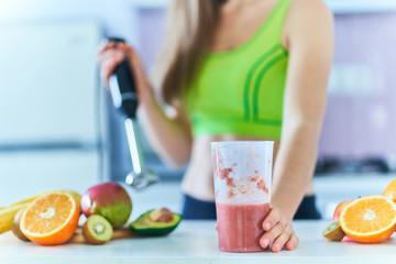 Obraz na płótnie Canvas Healthy fitness woman in sportswear prepares a fresh fruit smoothie using a hand blender at home in the kitchen. Vitamin diet drinks