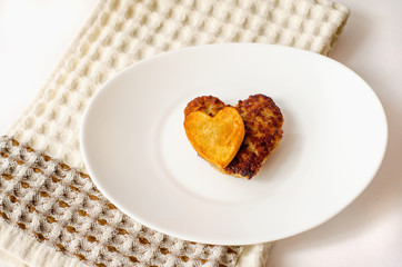 Fried cutlet in the shape of a heart, on it lies a heart of fried potatoes on a white oval plate. A gift for a loved one. Close up. Selective focus