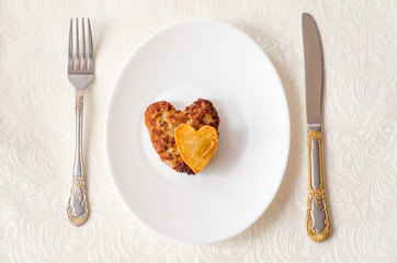 Fried cutlet in the shape of a heart, on it lies a heart of fried potatoes on a white oval plate with a fork and a knife. A gift for a loved one. Close up. Selective focus