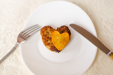 Fried cutlet in the shape of a heart, on it lies a heart of fried potatoes on a white oval plate with a fork and a knife. A gift for a loved one. Close up. Selective focus