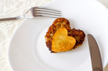 Fried cutlet in the shape of a heart, on it lies a heart of fried potatoes on a white oval plate with a fork and a knife. A gift for a loved one. Close up. Selective focus
