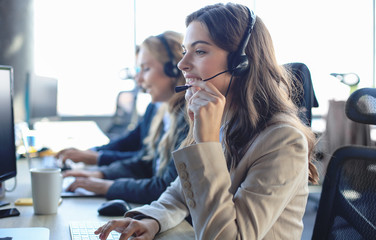 Female customer support operator with headset and smiling, with collegues at background. - obrazy, fototapety, plakaty