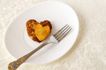 Fried cutlet in the shape of a heart, on it lies a heart of fried potatoes on a white oval plate with a fork and a knife. A gift for a loved one. Close up. Selective focus