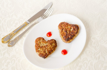 Two heart-shaped fried cutlets on an oval white plate lie on the table on a cream-colored kitchen napkin, along with a knife and fork. Surprise for a loved one for the holiday. Top view. Close up.