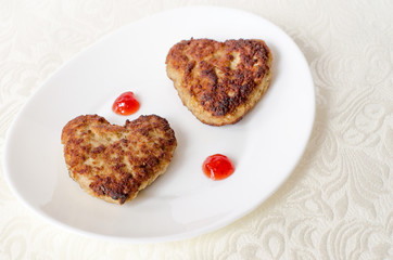 Two heart-shaped fried cutlets on an oval white plate lie on the table on a cream-colored kitchen napkin. Surprise for a loved one for the holiday. Top view. Close up.