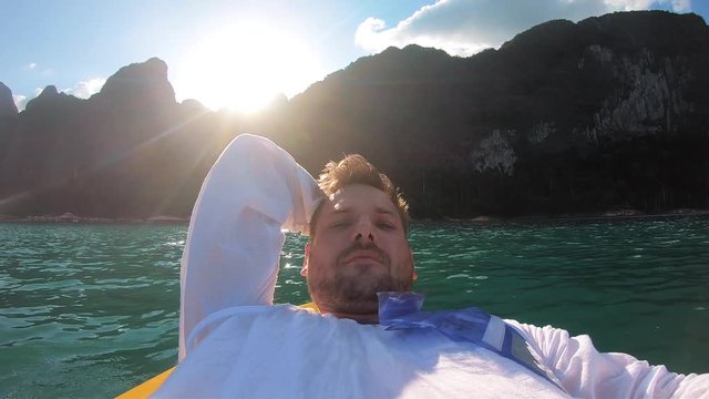 The blogger guy is lying on a kayak on the lake and takes selfie for social networks on Instagram,a beautiful natural landscape behind him,mountains,the sun,wildlife and the jungle.A man travels