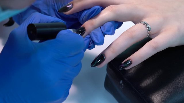Closeup view of beautiful manicured nails of woman and professional manicurist applying transparent finish top polish laer of gel polish. Black design with shiny sparkling cat eye effect. 4k video.