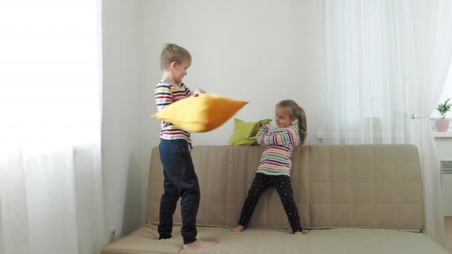 Brother and baby sister fighting by pillows on the sofa, slow motion