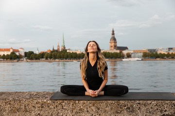 Yoga meditation and wellness lifestyle concept. Fit slim woman practicing yoga exercises near river Daugava and Vecriga old town background at sunset - Full sitting shot and meditating on rock beach