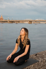 Fototapeta na wymiar Yoga meditation and wellness lifestyle concept. Fit slim woman practicing yoga exercises near river Daugava and Vecriga old town background at sunset - Full sitting shot and meditating on rock beach