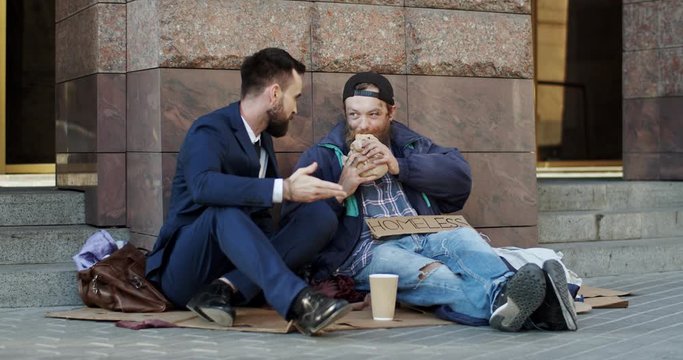 Rich Caucasian man in business style and homeless man beggar sitting together on the ground and talking cheerfully while man eating sandwich which businessman bought him.
