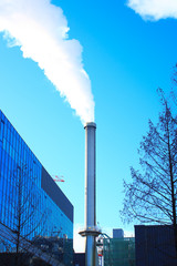Close-up on an industrial chimney. Polluting smoke in urban areas. Clear sky in the background. Building and vegetation without foliage in the foreground.