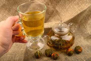 Hand holding a glass of tea, Flower tea brewed in a glass teapot and balls of flower tea on a background of rough homespun fabric. Close up.