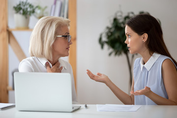 Middle-aged and millennial female colleagues arguing seated at workplace