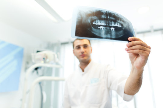 Young doctor dentist man in white uniform standing and looking at tooth picture in dental office in clinic with equipment at background, selective focus. Dental health and medical care concept