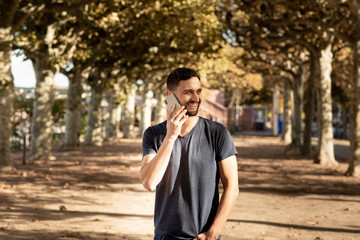 Young man in tshirt standing on street, speaks on a mobile phone and smiles. In the background is a tree lane. Frankfurt, Germany