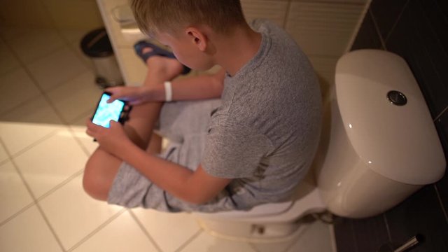 Young kid hiding in bathroom from parents to have time for playing online video games at his smartphone. Dressed boy sitting on seat of toilet and using blurry cell phone. Shallow depth of field.