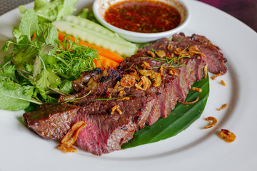 Slice medium rare grilled beef steak on top with fried garlic with Thai spicy sauce and vegetable on side.