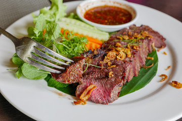 Using fork to stab and pick a piece of slice medium rare grilled beef steak on top with fried garlic with Thai spicy sauce and vegetable on side.