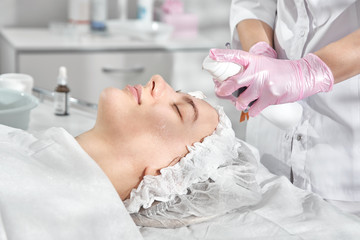 Professional beautician in salon provides comprehensive facial skin care for young woman. Pore cleansing procedures, all-season peeling, firming anti-aging mask