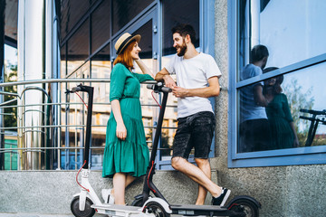 A young woman in dress and man talking to each other and enjoy a convenient way to travel around the city on electric scooters