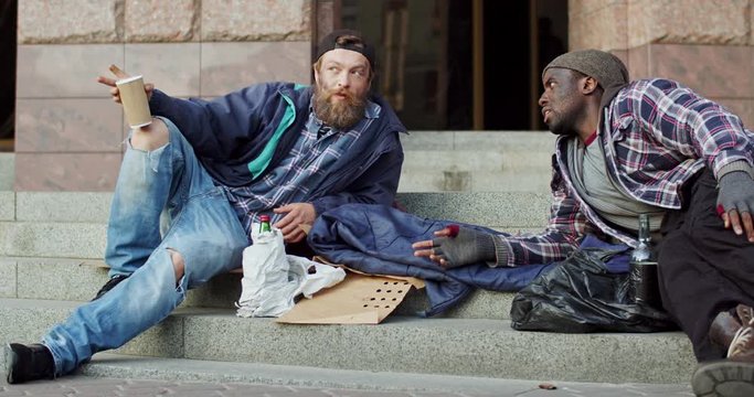 Caucasian and African American two homeless men beggars sitting on steps at the street and talking about their hard life.