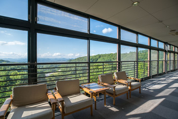 High level interior corridor in a modern building with floor-to-ceiling windows and natural scenery outside the window, some wooden chairs and table in front