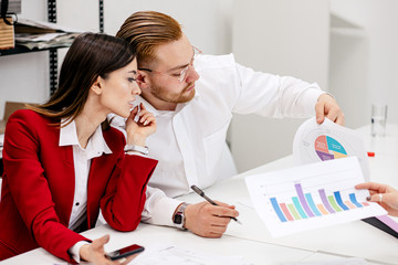 young caucasian business people discuss project together, sitting in modern white office wearing formal clothes, brunette woman and blond man holding papers with diagrams