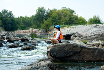 woman in the cap is sitting on the big stone above the white water river