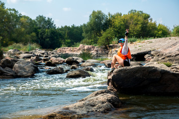woman in the cap is sitting on the big stone above the white water river