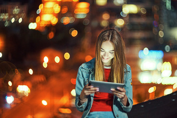 Girl on night cityscape background with street lights, using a digital tablet