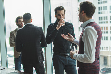 free time of caucasian colleagues at work in office, handsome men in formal wear stand next to panoramic window and have conversation. business people concept
