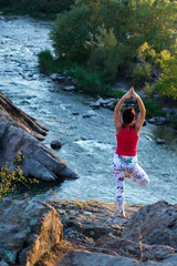 adult woman doing yoga asanas on the rocks above the white water river at sunset time