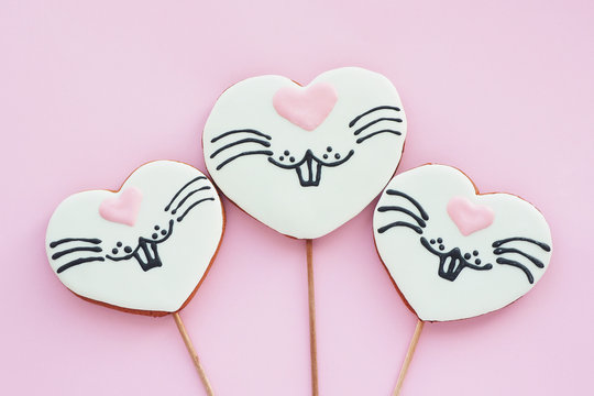.Three gingerbread cookies in the shape of a heart with a painted rabbit face on a stick on a pink background. Easter bunny concept..