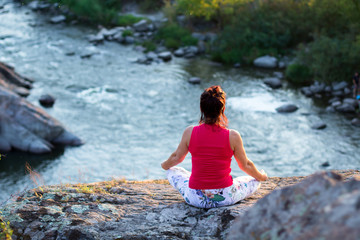 adult woman doing yoga asanas on the rocks above the white water river at sunset time