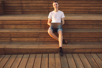 Portrait of a happy young male freelancer working on the street near the sea on a wooden platform with a laptop and smartphone on a sunny summer day. Remote work concept