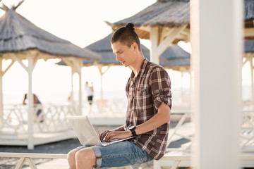 Portrait of a positive young remote worker guy with a laptop sitting under a straw canopy on the beach on a sunny summer day. Freelancer and outsourcing concept