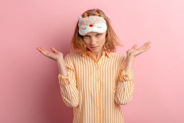portrait of young and beautiful caucasian woman before sleeping, want to sleep, stand shrugging isolated over pink background, wearing pajamas and blinfold for sleep