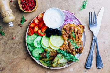 Lunch bowl with grilled chicken, red onion, tomato, cucumber, lemon, olive and sause on wooden background. Top view.