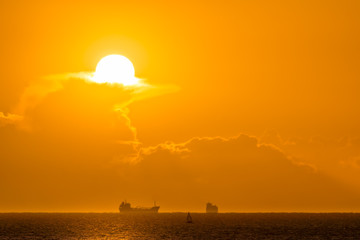 Fototapeta na wymiar Silhouette of cargo ships on far distance at golden hour. Sun, solar disc, shines from behind cloud. Bull Island, Dublin, Ireland