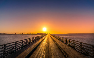 Long wooden bridge leading to Bull Island with silhouette of houses and lighthouse at golden hour. Sun, solar disc, just above horizon Dublin, Ireland