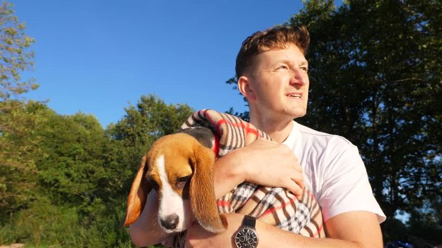 Handsome man care about sweet young beagle. Owner embrace puppy wrapped in plaid and pat head, try to warm him after swimming in cold sea. Green trees on background, low angle slow motion shot