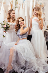 Naklejka premium three young romantic ladies in wedding dresses, future brides, going to be married, posing together at camera among dresses, holding flowers in hands
