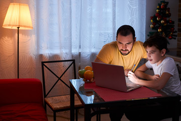 little caucasian boy sit doing homework in kitchen, using laptop, careful father support and help him to study, wearing casual clothes