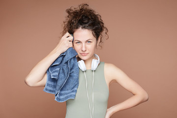 Shot of tender sensual female runner takes break, stands indoor with headphones around neck, has satisfied expression, wipes sweat with towel, isolated over beige studio wall. Sport, fitness concept.