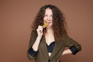 Fashionable excited curly haired blue eyed female biting plastic cart as being happy about...