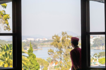 hotel bellboy in french colonial uniform looking out to view of alpine lake and garden in sunshine...