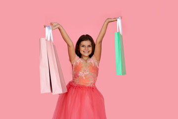 Pretty little girl wearing a pink dress is happy after sale and special offer shopping showing her colorful bags, over pink background.