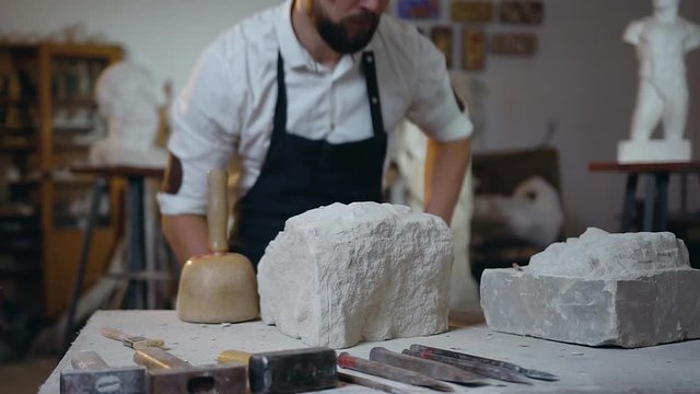Handsome creative bearded young statuary caming to the workplace and putting on glasses starting to work with white limestone using hammer and chisel